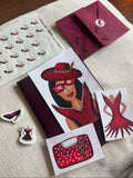A styled flat-lay of fashion-themed stationery and accessories on a cream fabric surface, including a printed card with an illustration of a woman in red, a matching dress illustration, a red polka-dot handbag drawing, a sheet of shoe-shaped stickers, and burgundy envelopes.