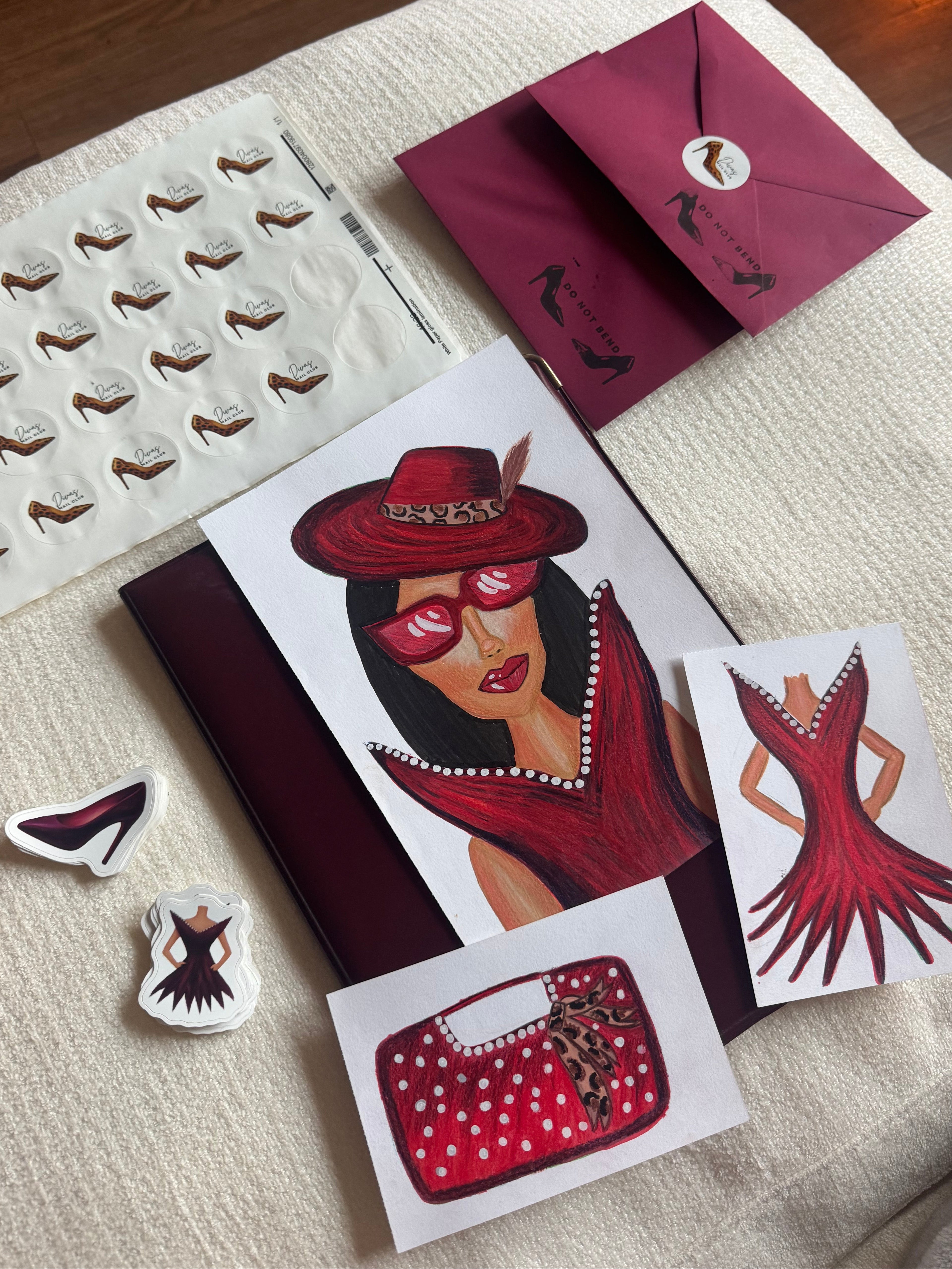 A styled flat-lay of fashion-themed stationery and accessories on a cream fabric surface, including a printed card with an illustration of a woman in red, a matching dress illustration, a red polka-dot handbag drawing, a sheet of shoe-shaped stickers, and burgundy envelopes.